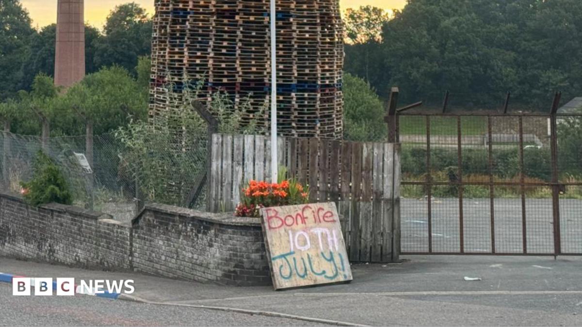 A bonfire of wooden pallets sits behind a fence, in front of the fence is a brick wall and a wooden sign is propped up against it with "bonfire 10th July" written on it