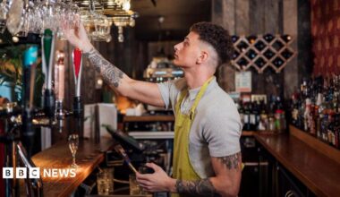 A bar staff member wearing a grey t-shirt and a yellow apron stands behind the bar holding a glass in his hand.