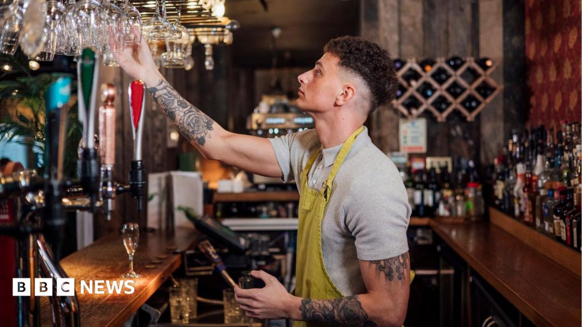 A bar staff member wearing a grey t-shirt and a yellow apron stands behind the bar holding a glass in his hand.
