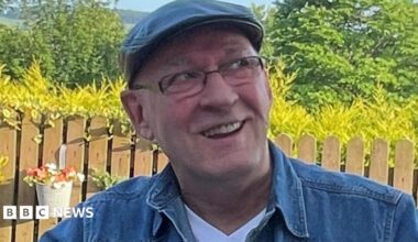 A man wearing a cap, a denim shirt and rectangular glasses sitting in a garden - he is smiling and looking off to the right of the camera