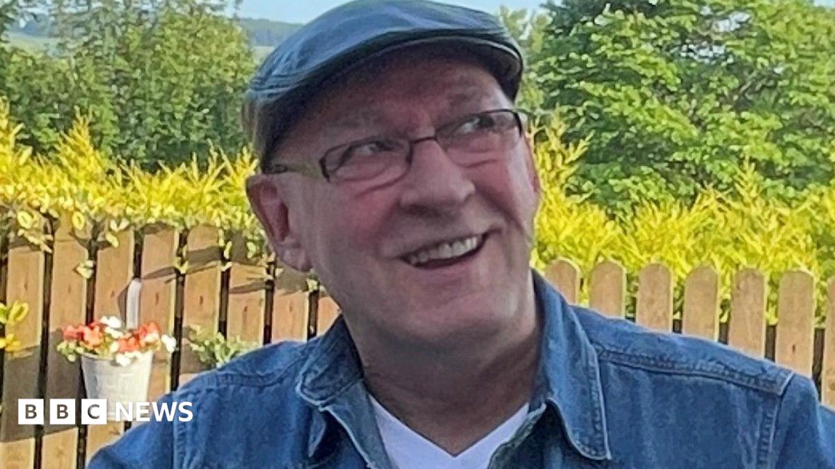 A man wearing a cap, a denim shirt and rectangular glasses sitting in a garden - he is smiling and looking off to the right of the camera