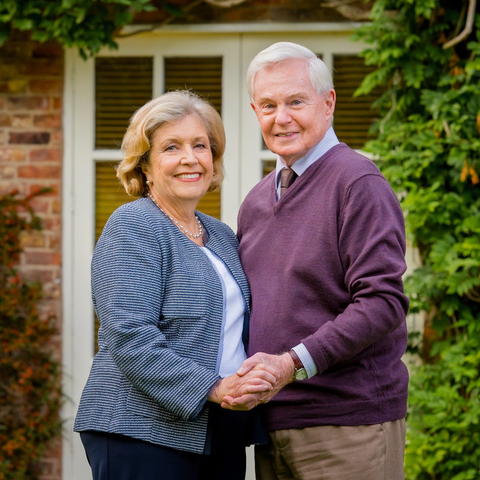 elderly couple smiling and holding hands outdoors with greenery.