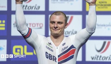 Matt Richardson in white British kit with GBR lettering raises arms aloft in front of sponsor background