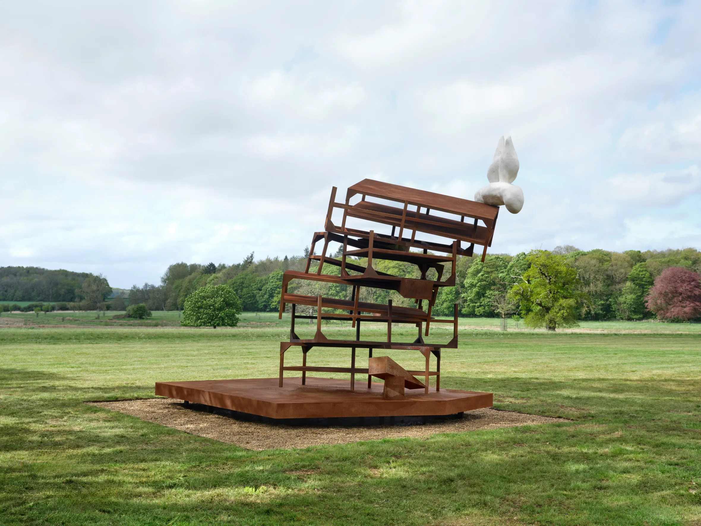 A metal sculpture resembling stacked, deconstructed chairs with a white abstract form on top, set on a gravel-covered base in a grassy field.