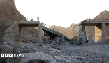 A damaged bridge with people standing on the remaining structure with mountains in the background, and rubble and rocks in the foreground.