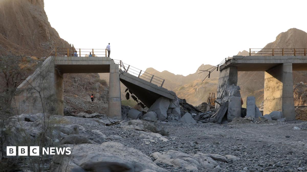A damaged bridge with people standing on the remaining structure with mountains in the background, and rubble and rocks in the foreground.