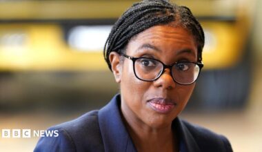 A picture of Kemi Badenoch from the shoulders up. She is wearing a dark blue suit jacket and black-rimmed glasses. Her hair, which is in braids, is tied back. She is looking to the left and the background is blurred.