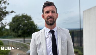 A smartly dressed man with dark hair and a beard wears a grey suit and black tie.