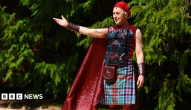 Alan Cumming, a man, is stood holding his arm out with trees behind him. He has a red beret on his head, a leather wrist band, a red cape, a tartan vest, tartan kilt, red knee high socks and a sporran which have the initials AC embroidered onto them. He is smirking slightly and looks as if he is welcoming people to something.