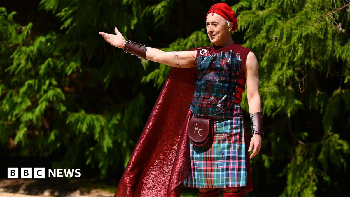 Alan Cumming, a man, is stood holding his arm out with trees behind him. He has a red beret on his head, a leather wrist band, a red cape, a tartan vest, tartan kilt, red knee high socks and a sporran which have the initials AC embroidered onto them. He is smirking slightly and looks as if he is welcoming people to something.
