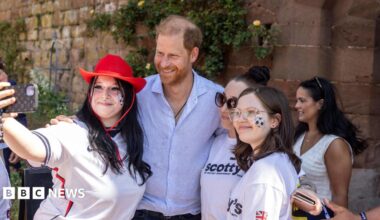 Prince Harry, wearing a light blue shirt, smiles for a photo with three young women. The woman taking the selfie is wearing an England football shirt and a red cowboy hat.