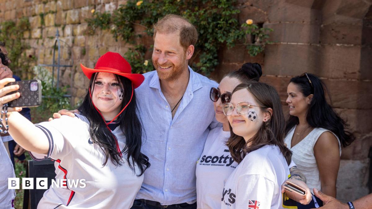 Prince Harry, wearing a light blue shirt, smiles for a photo with three young women. The woman taking the selfie is wearing an England football shirt and a red cowboy hat.