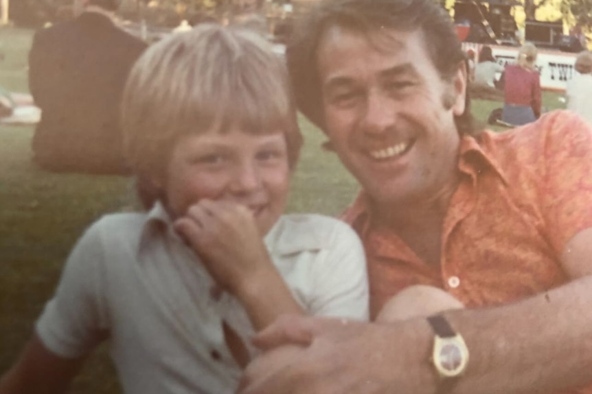 A old-style photo of a blonde haired boy in a white top with a brown haired man in an orange top.