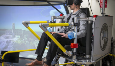 A brown and yellow platform holding a seat that is raised up in the air is holding one passenger. The passenger is wearing virtual reality goggles and a harness. There is a TV monitor to the right of the monitor showing the words “liftoff” and a second screen behind the passenger.