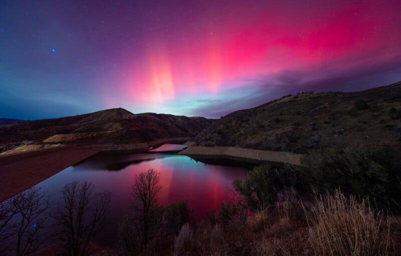A vibrant aurora displays pink, red, and purple lights in the night sky over a calm lake surrounded by hills and sparse vegetation, with reflections of the colors visible in the water.