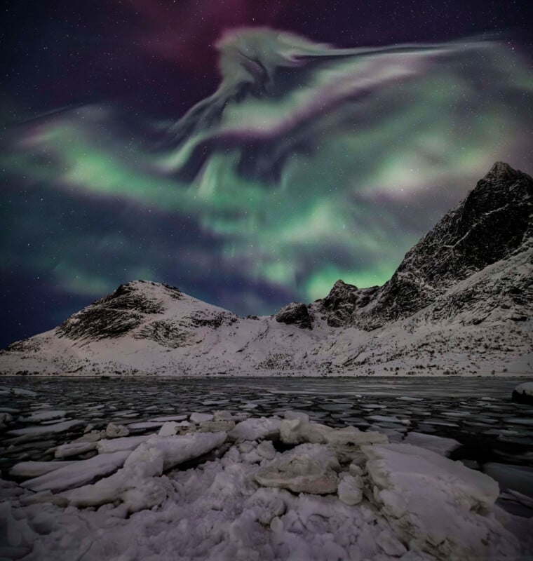 Green and purple aurora borealis lights swirl in the night sky above snowy mountains and a frozen, ice-covered landscape, creating a dramatic, natural scene.