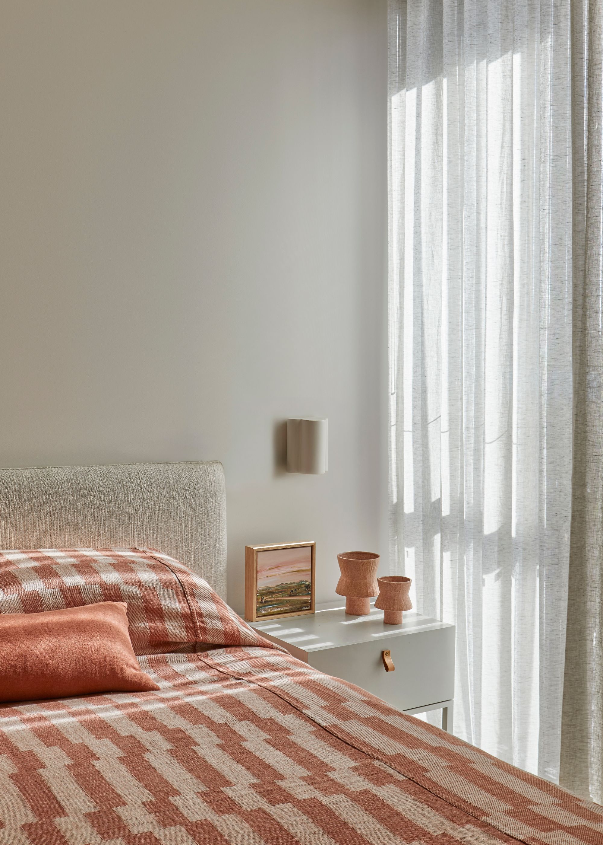 A bright bedroom with sheer curtains, a patterned duvet, a coral pillow, a white nightstand, a pair of vases, and a frame