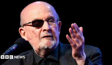 Rushdie speaking in March 2026 into a microphone. He has a darkened lens over his wounded eye