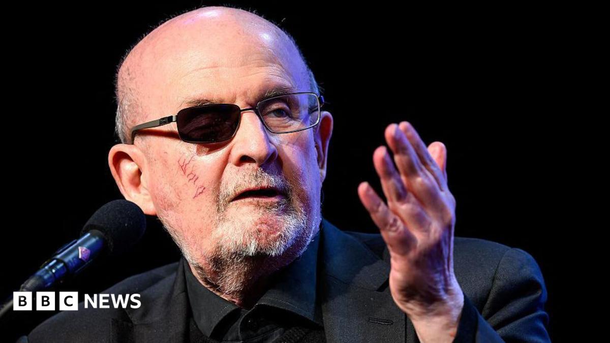 Rushdie speaking in March 2026 into a microphone. He has a darkened lens over his wounded eye