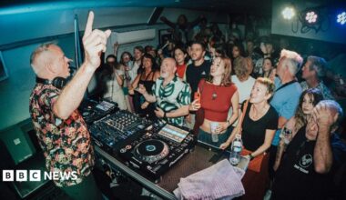 DJ Fatboy Slim points upward while performing behind a mixing deck in a compact room filled with a tightly packed audience, with people standing shoulder to shoulder, some holding drinks, under bright stage lights and low ceilings.