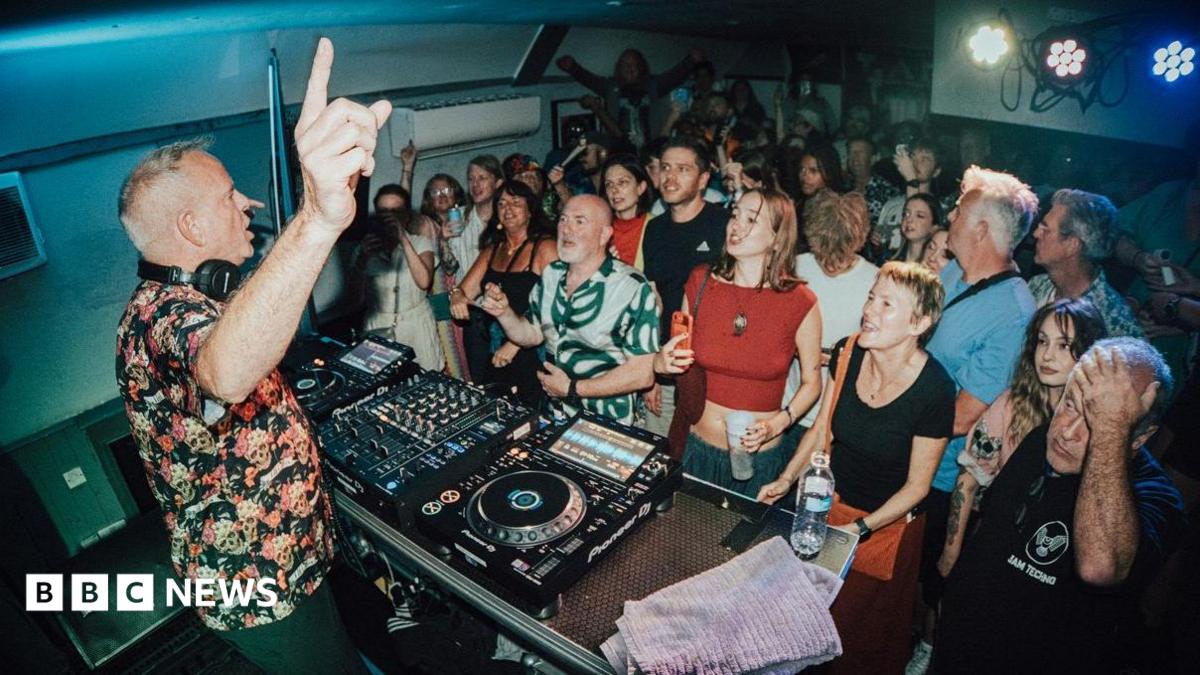 DJ Fatboy Slim points upward while performing behind a mixing deck in a compact room filled with a tightly packed audience, with people standing shoulder to shoulder, some holding drinks, under bright stage lights and low ceilings.