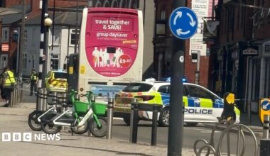 Police vehicles with flashing lights parked behind a bus at a town centre junction, with part of the road cordoned off, bicycles parked nearby and surrounding shops and buildings visible