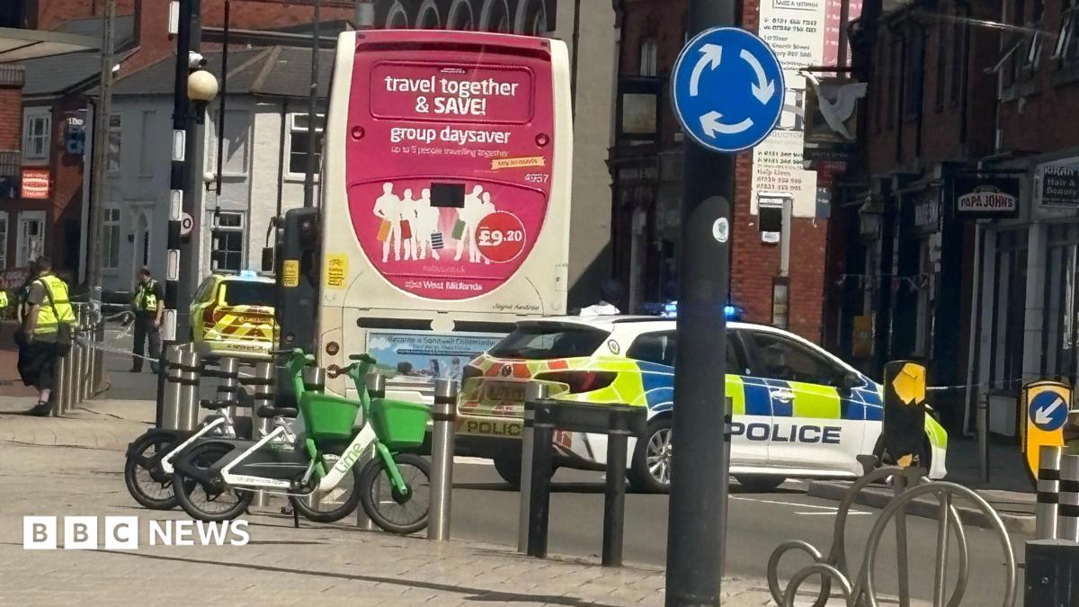 Police vehicles with flashing lights parked behind a bus at a town centre junction, with part of the road cordoned off, bicycles parked nearby and surrounding shops and buildings visible