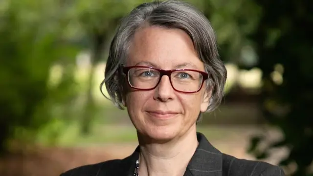 A woman with grey bobbed hair and black glasses wearing a dark blazer. She is outdoors with greenery behind her and smiling.