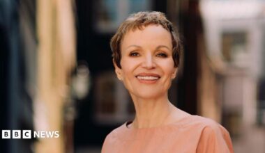 Marcia Kilgore with cropped short brown hair smiling at the camera with a blurred background wearing a nude coloured round neck top