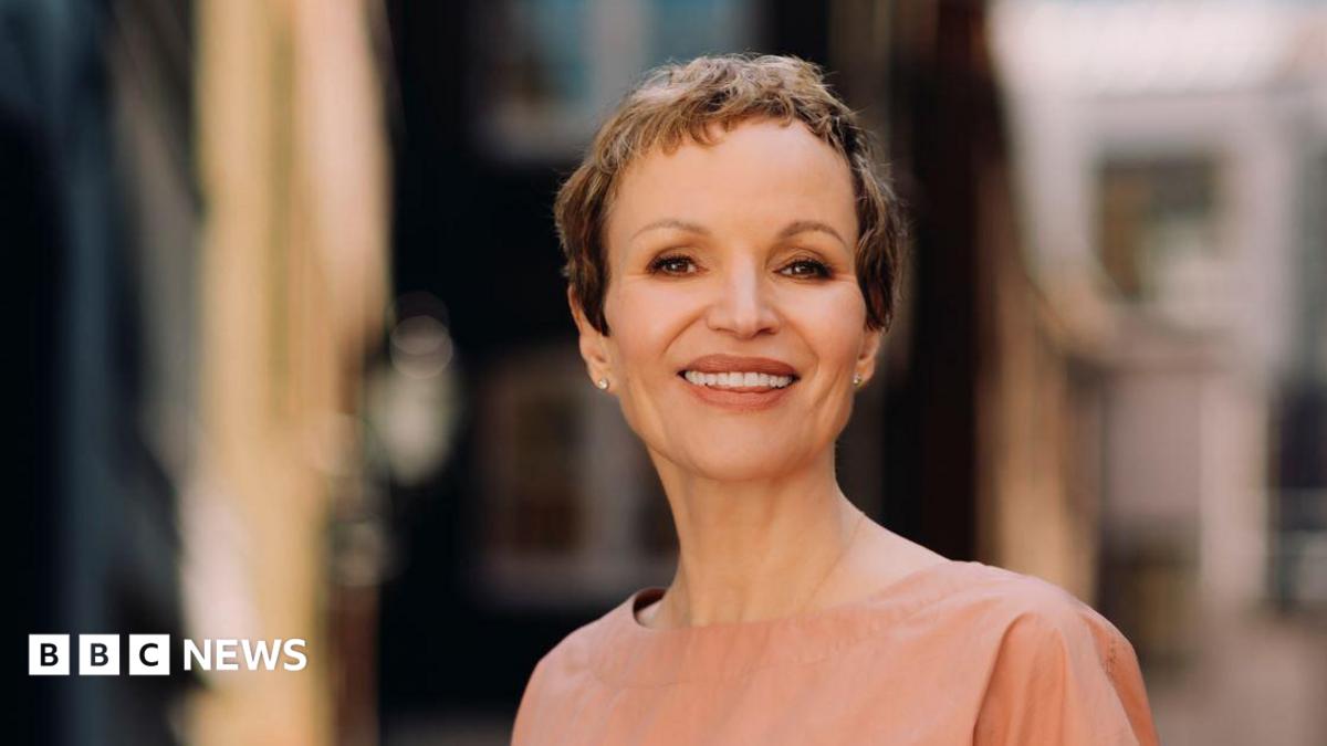 Marcia Kilgore with cropped short brown hair smiling at the camera with a blurred background wearing a nude coloured round neck top