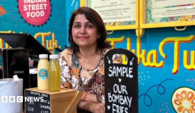 A woman with brown hair stood behind a food counter, the signs in the background are bright blue, with the word Tukka Tuk written in bright yellow. The woman is wearing an orange and white top.
