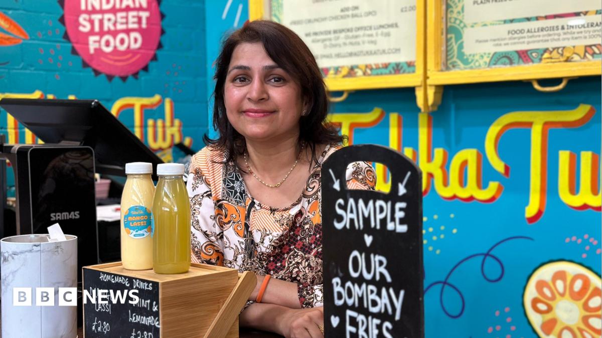 A woman with brown hair stood behind a food counter, the signs in the background are bright blue, with the word Tukka Tuk written in bright yellow. The woman is wearing an orange and white top.