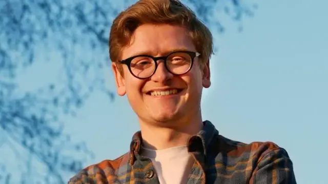 A close up of Richard. He has curly brown hair and is wearing a pair of glasses, a check shirt, and is smiling.