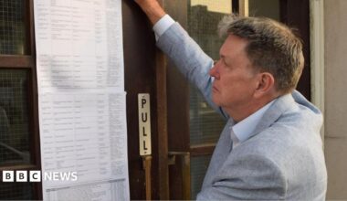 Ian Davidson posting the statement of persons nominated outside Clacton Town Hall. He is on the right, has short light brown hair and is wearing a light grey jacket over an open-neck shirt. His right arm is upraised as he puts the long list of typed names on to the brown wooden and glass door.