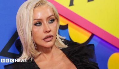 Christina Aguilera at The Abbey 35th Anniversary Party held at The Abbey on May 20, 2026 in Los Angeles, California. She is wearing a black top and has shoulder length blonde hair. Christina is standing in front of a brightly coloured blue, yellow and pink backdrop.