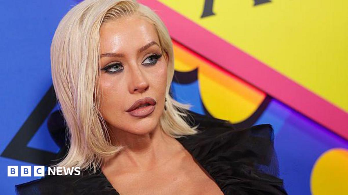 Christina Aguilera at The Abbey 35th Anniversary Party held at The Abbey on May 20, 2026 in Los Angeles, California. She is wearing a black top and has shoulder length blonde hair. Christina is standing in front of a brightly coloured blue, yellow and pink backdrop.