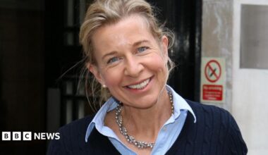 Katie Hopkins smiles at the camera with her head tilted to her right shoulder. She is wearing a navy blue jumper with a blue shirt underneath.