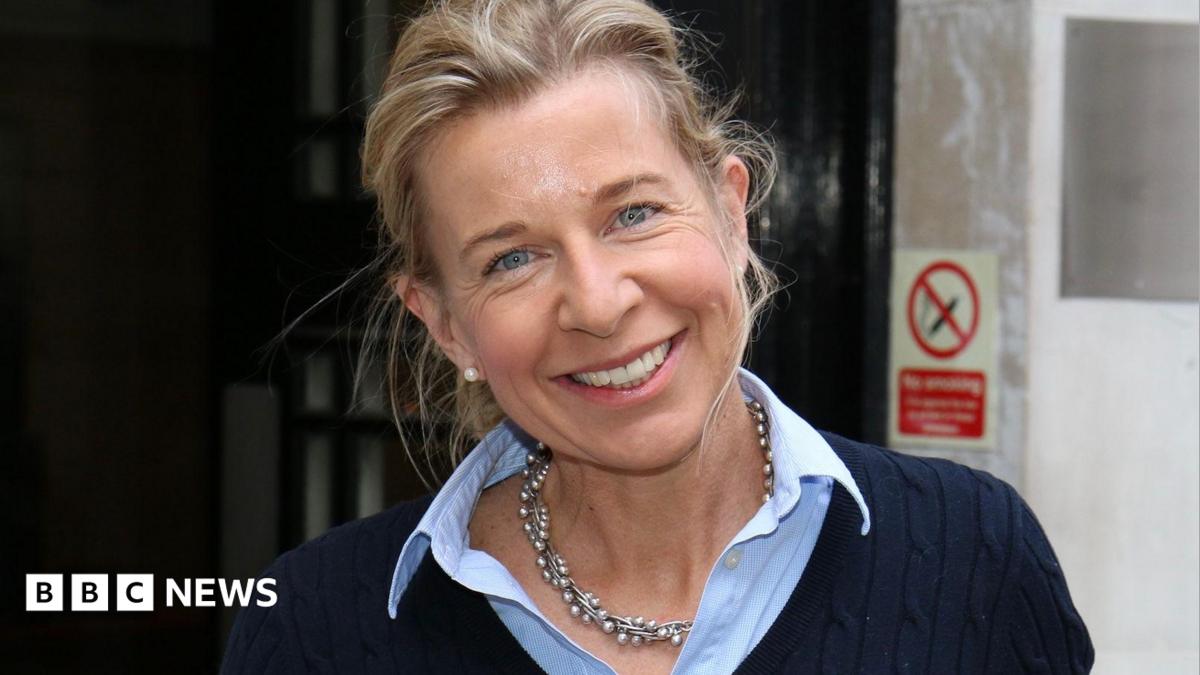 Katie Hopkins smiles at the camera with her head tilted to her right shoulder. She is wearing a navy blue jumper with a blue shirt underneath.