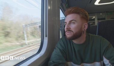 Wegrzyn has a ginger quiff and beard and looks out of a train window as it moves down the tracks, deep in thought. They have two black earrings in their ears and are wearing a green and grey jumper.