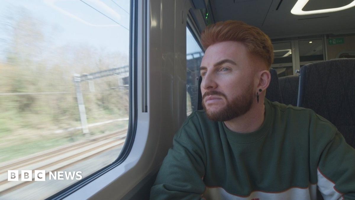 Wegrzyn has a ginger quiff and beard and looks out of a train window as it moves down the tracks, deep in thought. They have two black earrings in their ears and are wearing a green and grey jumper.