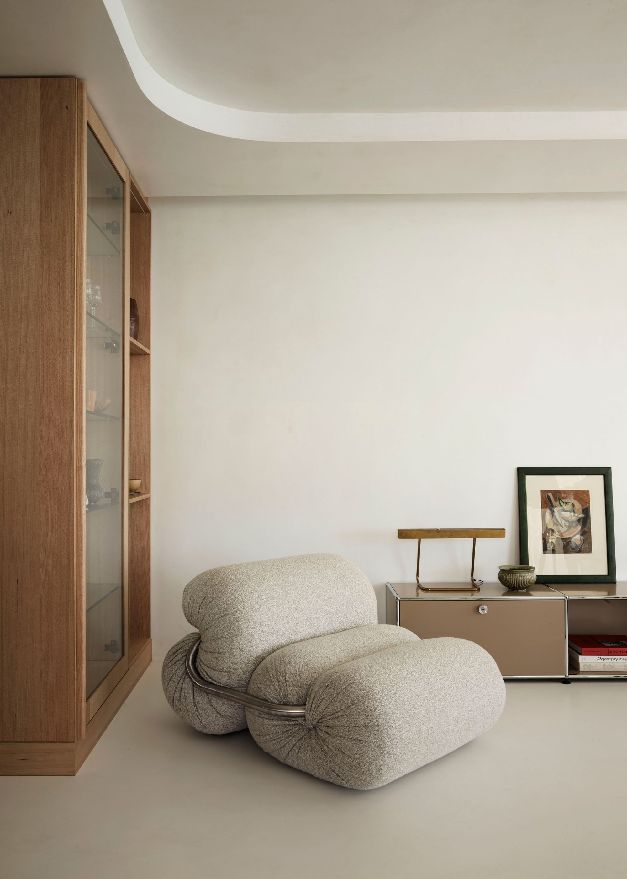 A living room with a glass display cabinet, a gray structured sofa chair, a light brown cabinet with a lamp, a bowl, and a leaning painting