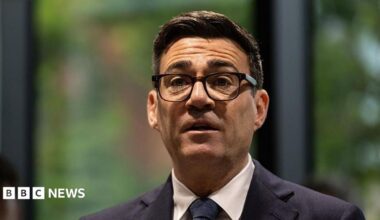 A picture of Andy Burnham from the shoulders up. He is wearing a dark suit with a white shirt and dark tie, has thick-rimmed black glasses on, and has short, dark brown hair. The background behind him is blurred and he is looking forward with his mouth slightly open and his eyebrows raised.