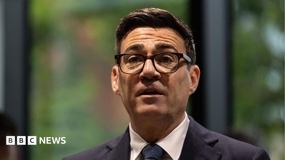 A picture of Andy Burnham from the shoulders up. He is wearing a dark suit with a white shirt and dark tie, has thick-rimmed black glasses on, and has short, dark brown hair. The background behind him is blurred and he is looking forward with his mouth slightly open and his eyebrows raised.