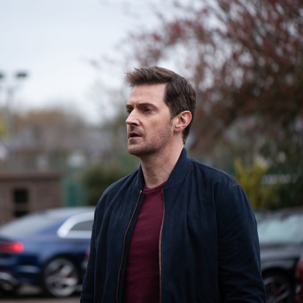 man in a dark jacket standing outdoors in front of a blurred background with trees and a car.