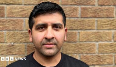 A picture of former cricketer Majid Haq. He is wearing a black T-shirt and is leaning against a brick wall, looking into the camera lens.
