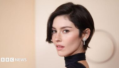 Gracie Abrams poses on the red carpet of the 2026 Oscars, in a black dress with a choker, and sparkling diamond earrings.