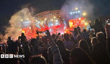 A large crowd scene at night with hundreds of music fans facing a large stage lit by pink lighting