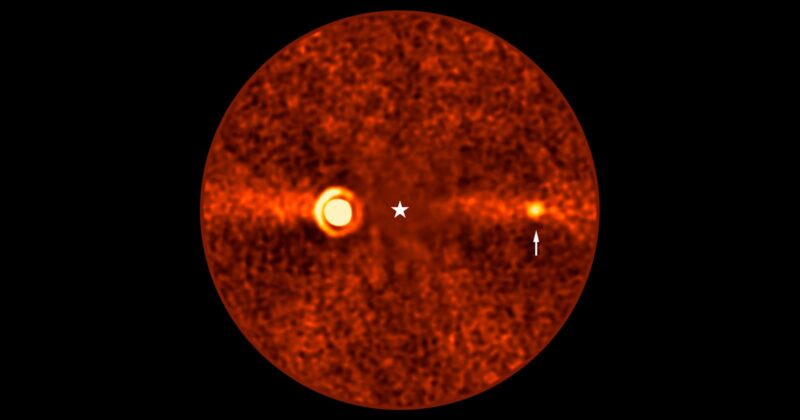 Orange-hued, grainy circular image of a star at the center with a glowing ring to the left and a smaller bright spot to the right, indicated by an arrow. The background is dark with scattered noise.