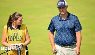 A golfer in a navy polo shirt and cap strides up the fairway of a golf course next to his female caddie.