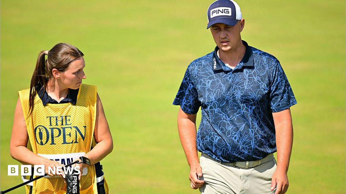 A golfer in a navy polo shirt and cap strides up the fairway of a golf course next to his female caddie.